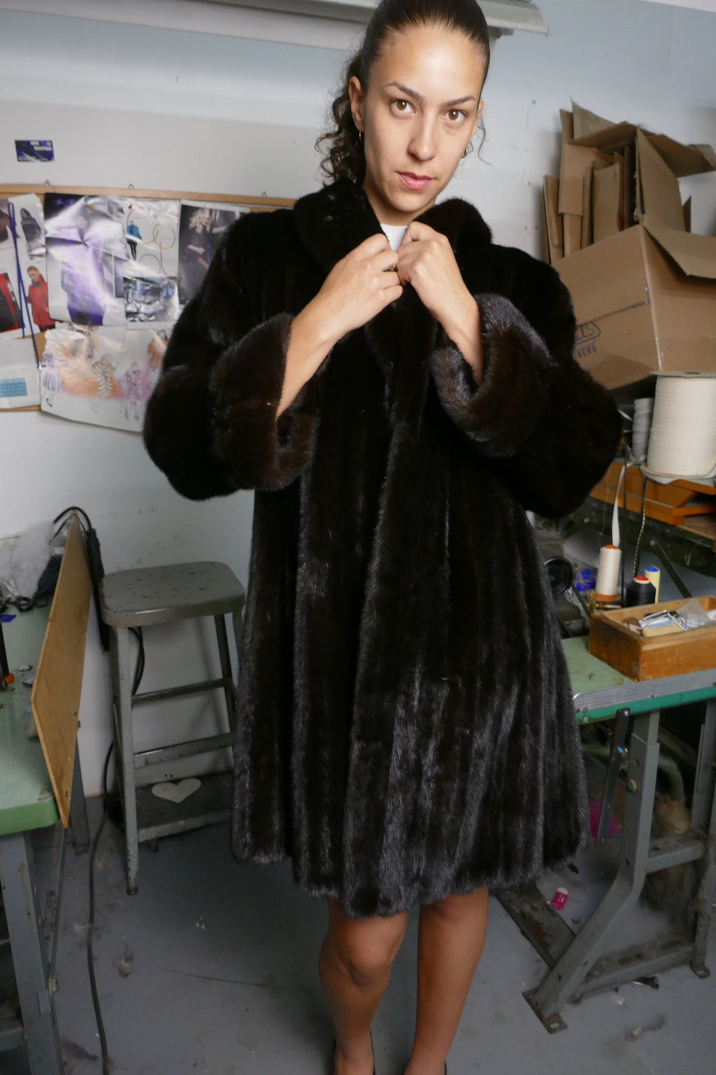 A woman wearing a dark brown plush fur coat with a wide collar, standing in a cluttered workshop or studio environment.