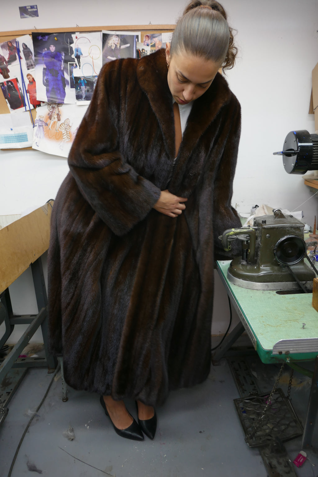A woman wearing a long, luxurious dark brown fur coat stands in what appears to be a tailoring or fur workshop, looking down while fastening the coat. The background features sewing machines, a workbench, and sketches pinned on a board.