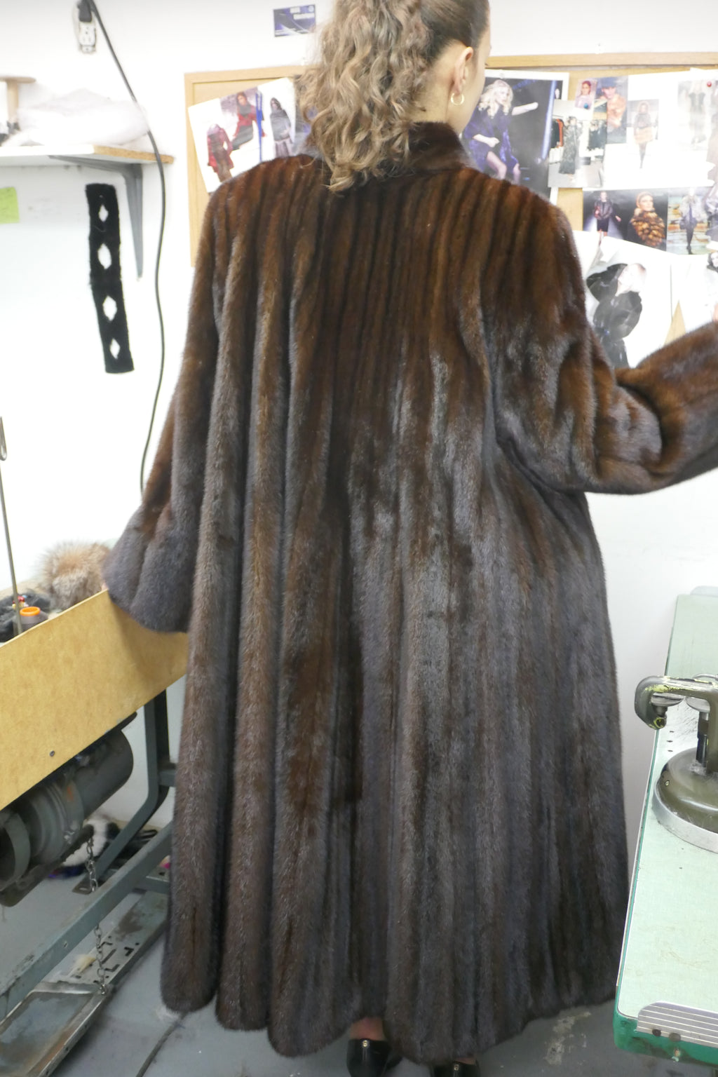 A person viewed from behind wearing a long, dark brown fur coat in what appears to be a workshop or studio interior.