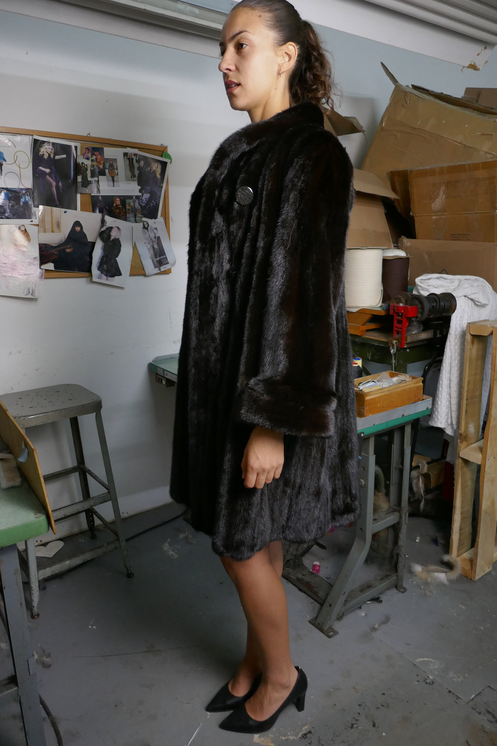A woman wearing a dark brown fur coat and black heels, standing in a workshop or studio environment with tools, cardboard boxes, and pinned photos on the wall.
