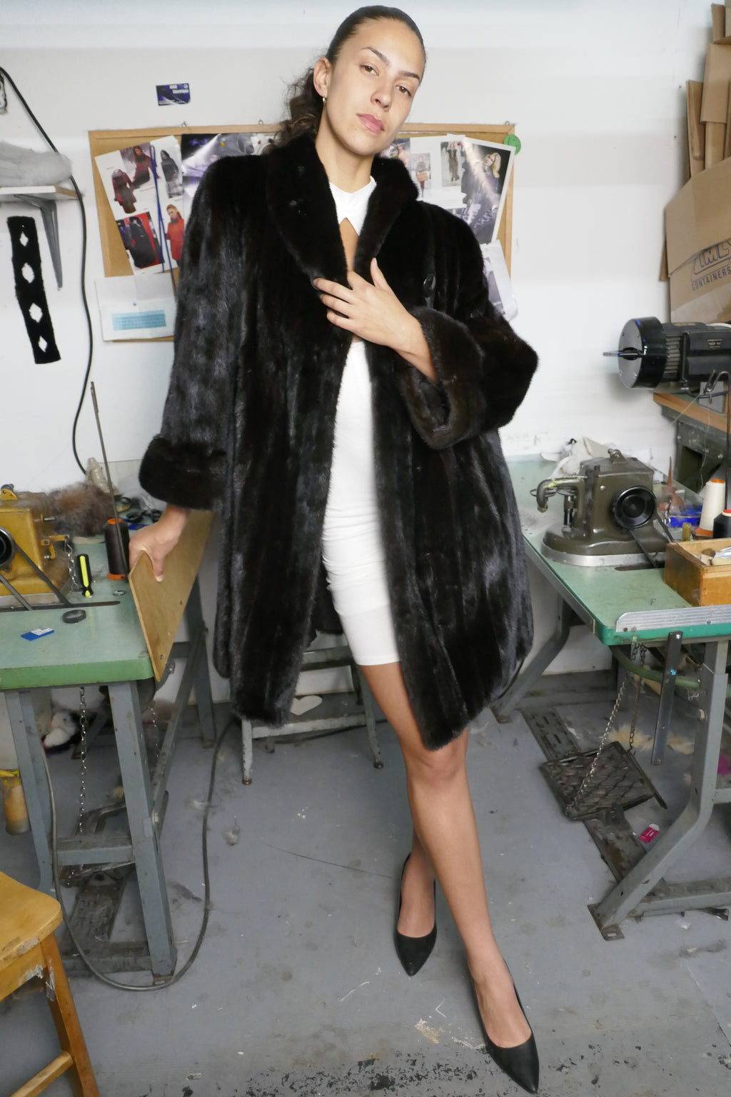 A woman wearing a long black fur coat over a white dress and black heels, standing in a workshop with sewing tables and equipment in the background.