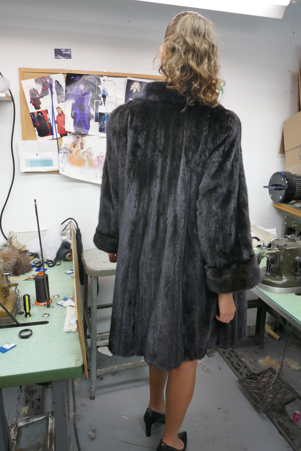 A woman viewed from behind wearing a dark gray faux fur coat in what appears to be a tailoring or sewing workshop.