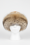 A large plush fur hat with a natural spotted pattern in beige, brown, and cream tones, displayed on a white mannequin head against a light gray studio background.